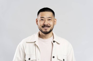 Gülümseyen Koreli erkek gri arka planda izole edilmiş kameraya bakıyor. Yakışıklı genç adam stüdyoda duruyor, resim için poz veriyor.