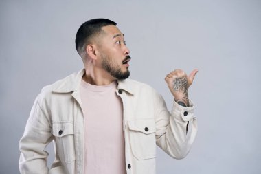 Şaşırmış Asyalı sakallı adam tek eliyle fotokopi alanını gösteriyor ve fikir sunumu için boş duvar gösteriyor. Kapalı alanda çekilen çekim gri arka planda izole edildi 