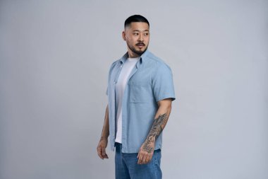 Tarz sahibi Asyalı yakışıklı bir adamın portresi. Sıradan kıyafetler giyiyor, kolunda dövmesi var. Gri arka planda izole bir şekilde başka tarafa bakıyor. Boşluğu kopyala