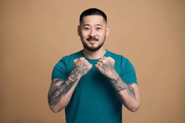 Yakışıklı Asyalı sakallı bir adamın portresi. Sıradan kıyafetler giyiyor. Dövmeli kolları var ve pozitifmiş gibi poz veriyor. Kahverengi arkaplanda izole 