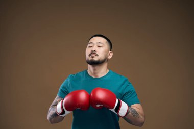 Motive olmuş yeşil tişörtlü Asyalı adam ve kırmızı boks eldivenleri kahverengi arka planda, gözleri kapalı ve rahatlatıcı. 