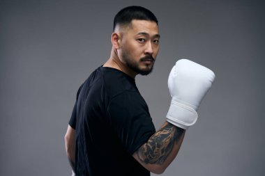 Siyah tişörtlü ciddi bir Koreli boks eldiveni takmış kolları dövmeli ve gülümsemeden kameraya dikkatlice bakıyor. Spor ve motivasyon kavramı 