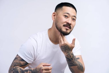 Yüzüne krem sürerek beyaz tişört giyen neşeli genç sakallı adam. Asyalı adam ileriye bakıyor ve gülümsüyor. Beyaz arkaplanda izole 