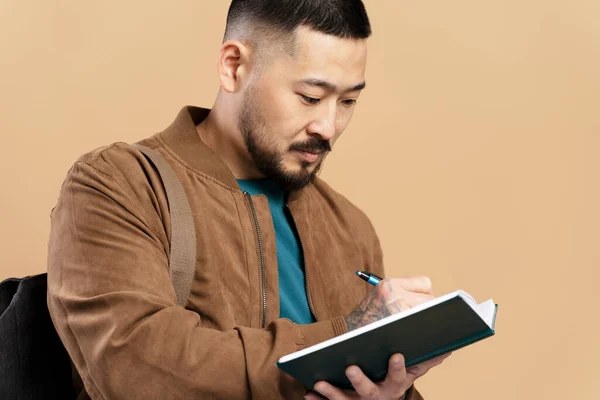 严重的亚洲学生写在笔记本与袋隔离米色背景 穿着棕色夹克的迷人男人 — 图库照片