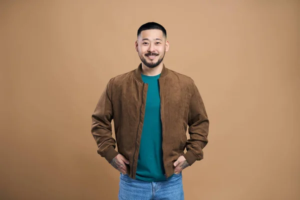 笑顔でカメラを見て 幸せな顔でポーズを取るカジュアルな服を着てハンサムな韓国髭の男の肖像画 茶色の背景に隔離された屋内スタジオショット — ストック写真