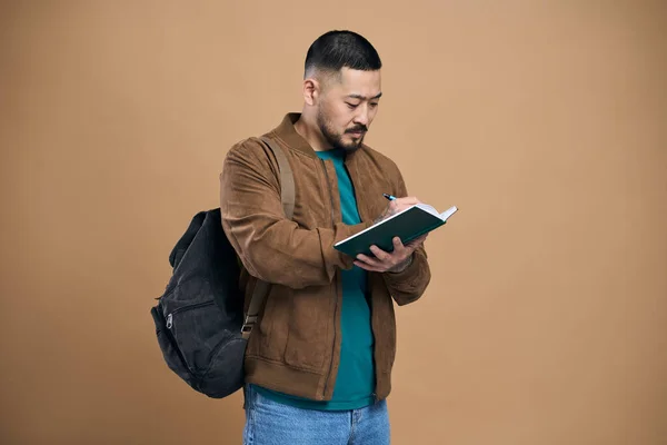 Asian man journalist in jacket making notes in paper notebook, writing business idea, future plans, checking appointment in schedule diary. Indoor studio shot isolated on brown background