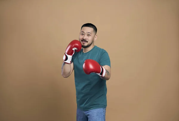 Positive asian man in green t shirt wearing red boxing gloves standing in fight pose and smiling to camera. Isolated over brown wall background