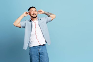 Güzel dövmeli, kulaklık takan, mavi arka planda izole edilmiş müzik dinleyen yakışıklı Asyalı bir adam. Teknoloji, olumlu duygu kavramı
