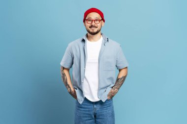 Çekici, gülümseyen Asyalı, stil dövmeli, mavi arka planda izole edilmiş, kopyalama alanı olan. Kırmızı şapkalı, mavi kotlu, gözlüklü, kameraya bakan kendine güvenen mutlu Koreli hippi.