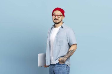 Sırıtkan zeki Asyalı öğrenci dizüstü bilgisayarını mavi arka planda, eğitim konseptinde izole etmiş. Şık dövmeli, kırmızı şapkalı, kameraya bakan gözlüklü Koreli serbest yazar portresi.