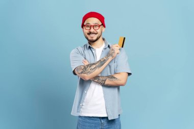 Başarılı gülümseyen, kırmızı şapkalı, şık gözlüklü, elinde kredi kartıyla mavi arka planda izole edilmiş kameraya bakan Asyalı bir adam. Kendine güveni tam mutlu serbest çalışan ödeme alır. Alışveriş, satış konsepti.