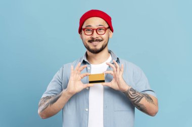 Mavi arka planda kredi kartı tutan yakışıklı, gülümseyen Asyalı adamın portresi.