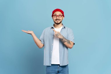 Yakışıklı gülümseyen Asyalı adam şık gözlükler takıyor, kırmızı şapka takıyor, el ele tutuşuyor, fotokopi alanını işaret ediyor. Alışveriş, satış konsepti.