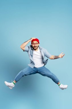 Yakışıklı heyecanlı Asyalı adam yüksekten atlıyor, mavi arka planda izole edilmiş eğleniyor. Kırmızı şapkalı, şık gözlüklü, beyaz ayakkabılı duygusal Koreli dansçının portresi. Mağaza, alışveriş, satış konsepti