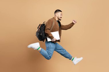 Sırt çantalı, dizüstü bilgisayarı olan, hızlı koşan meşgul duygusal Asyalı öğrencinin portresi. Bej arka planda izole edilmiş üniversiteye gitmek için acele et. Eğitim kavramı 