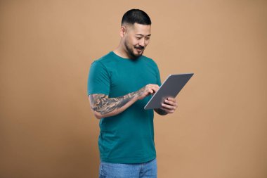 Kendine güvenen Asyalı adam elinde modern dijital tabletle videolar izliyor. İletişim bej arka planda izole edilmiş. Mutlu Koreli hipster internetten alışveriş yapıyor, bir şeylere bakıyor, yemek ısmarlıyor.                            