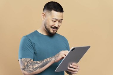 Yakışıklı, gülümseyen Asyalı adam elinde dijital tabletle videolar izliyor. İletişim bej arka planda izole edilmiş. Mutlu Koreli hipster internet üzerinden alışveriş yapıyor, web sitesinden yemek sipariş ediyor. Teknoloji konsepti