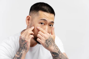 Yakışıklı Asyalı adamın sivilceleri temizleyen, beyaz arka planda izole edilmiş aynaya bakan portresi. Güzellik, cilt bakımı, sabah rutini konsepti 