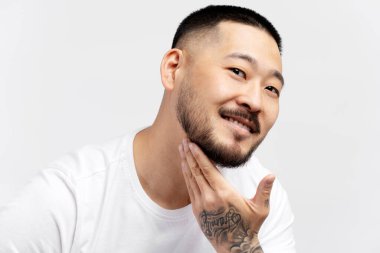 Yakışıklı, gülümseyen Asyalı adam yüzüne dokunuyor, düzgün sakallı beyaz aynaya bakıyor.