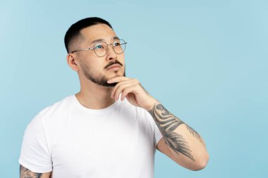 Yakışıklı dalgın Asyalı adam, yüzünün yanında el ele tutuşup mavi arka planda izole edilmiş yaratıcı bir çözüm arıyor. Genç ciddi bir üniversite öğrencisinin portresi. Başka tarafa bakıyor, düşünüyor. Eğitim kavramı