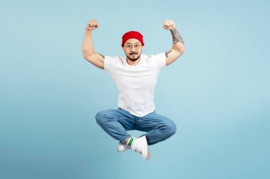 Güçlü Asyalı bir adam, vücut geliştiren, stil dövmeli, mavi arka planda izole olmuş kasları olan. Koreli kaslı erkek lotus pozisyonunda yükseğe zıplıyor, kameraya bakıyor. Spor, spor, sağlıklı yaşam tarzı.
