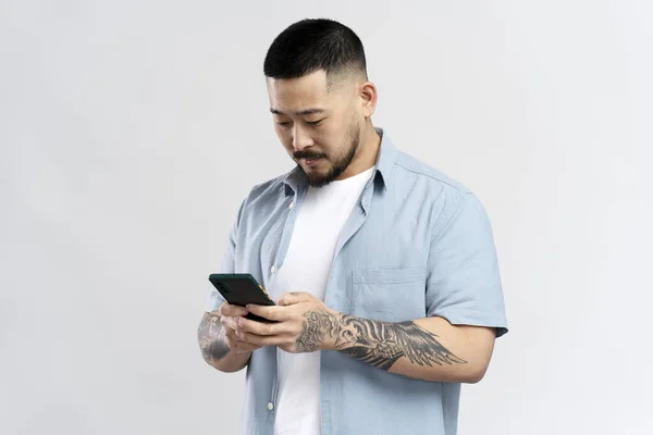 英俊的亚裔男子 有时髦的纹身 手持手机阅读短信 在白色背景下与外界隔绝 韩国嬉皮士家伙使用移动应用 网上购物 订购食物 — 图库照片