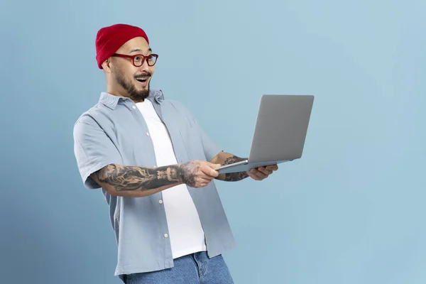 Excited stylish asian man holding laptop computer, watching videos with 5g high speed internet connection isolated on blue background. Shocked hipster guy shopping online with sales. Modern technology