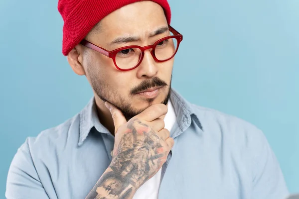 英俊的 沉思的 头戴红帽子的亚洲男子的肖像 蓝色背景的时髦眼镜 严重的韩国嬉皮士头脑风暴 计划在室内启动 工作室拍摄 — 图库照片
