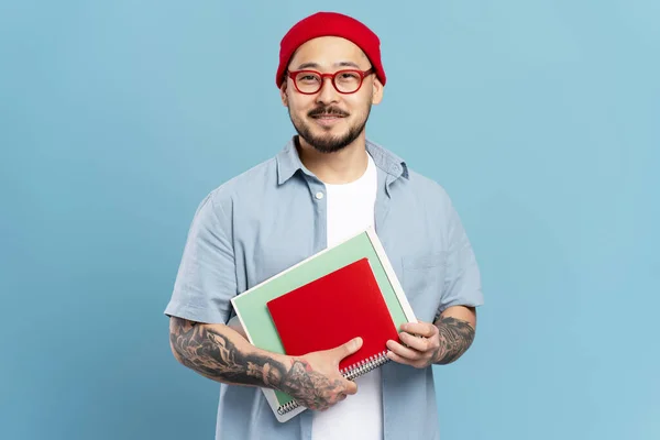 俊俏的笑脸 聪明的亚洲学生 拿着蓝色背景 教育观念孤立的书籍 韩国自由撰稿人的肖像 纹身时髦 头戴红帽子 目镜看着相机 — 图库照片