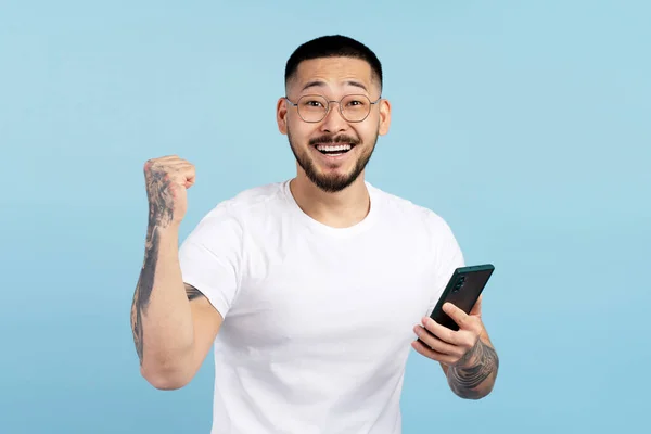 モバイルゲームをプレイするスマートフォンを保持している大喜びのアジア人男性 ブルーの背景で孤立したお祝いの成功 興奮した韓国のギャンブラースポーツ賭博 お金を稼ぐ 販売とオンラインで幸せな男性のショッピング — ストック写真