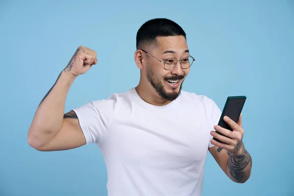 Overjoyed asian man holding mobile phone sports betting, win money isolated on blue background. Emotional asian gambler playing mobile game celebration success