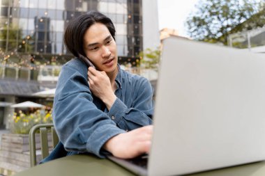 Genç Japon iş adamı dizüstü bilgisayar kullanıyor, cep telefonuyla konuşuyor, açık havada çalışıyor. İş yerinde oturan yakışıklı Asyalı adam. Uzak meslek, teknoloji konsepti