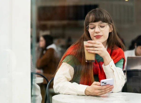 Cute young woman in stylish eyeglasses drinking coffee, holding mobile phone. Attractive female online ordering, technology concept