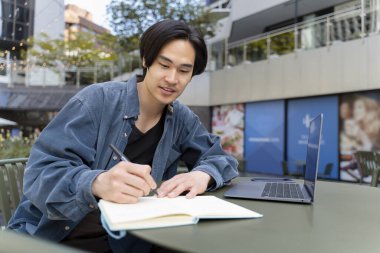 Düşünceli yakışıklı Japon öğrenci öğrenim görüyor, dil öğreniyor, online eğitim kavramı geliştiriyor. Asyalı, serbest çalışan, dizüstü bilgisayar kullanan, not alan, açık havada proje yapan biri. 