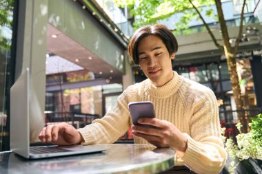 Yakışıklı gülümseyen Asyalı adam cep telefonu tutuyor, e-postaları kontrol ediyor, mesajları okuyor. Genç kendine güvenen Japon işadamı, modern kafede online olarak çalışırken dizüstü bilgisayar kullanıyor. Teknoloji konsepti 