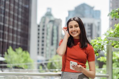 Esmer, gülümseyen Hintli, cep telefonu tutan ve şehir sokağında kameraya bakan bir öğrenci. Mutlu Asyalı kadın dışarıda mobil uygulama alışverişi yapıyor. Mobil bankacılık, teknoloji 