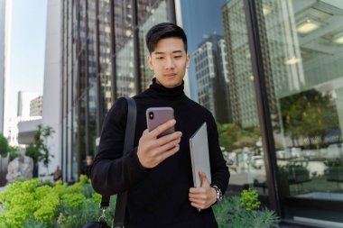 Laptop tutan gülümseyen Asyalı adamın portresi, açık havada cep telefonundan postaları kontrol ediyor. Şehir caddesinde çantasıyla dolaşan yakışıklı Çinli öğrenci. İnternetten alışveriş yapıyor.