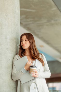 Profesyonel, güzel iş kadını elinde dizüstü bilgisayar ve içki bardağıyla modern ofise bakıyor. Kahve içen çekici finansör, mola. İş konsepti