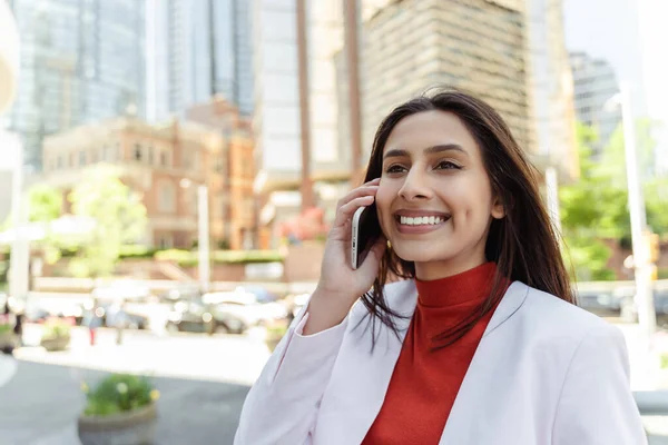 Authentic portrait of beautiful confident Indian woman talking on mobile phone, answering call looking away standing on urban street. Technology concept