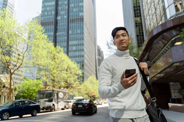 Attractive asian tourist standing on city street holding mobile phone looking away. Smiling chinese businessman using smartphone outdoors in city. Concept of travel, business