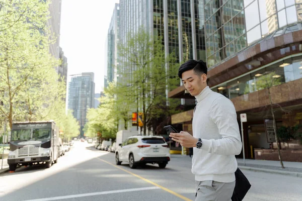 Portrait of positive asian man looking at mobile phone walking on city street. Attractive chinese student chatting, online shopping
