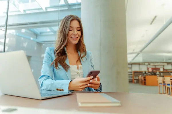 Portrait of smiling woman in formal wear holding mobile phone using laptop, sitting at workplace in office, library, remote work, online shopping. Female student studying online education