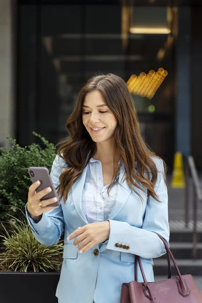 Smiling woman wearing blue jacket, hairstyle, holding mobile phone, text message, checking mail on the street. Confident businesswoman receive payment. Concept of successful business lifestyle