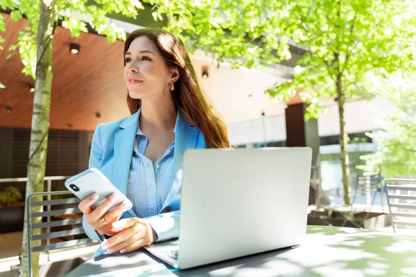 Attractive female freelancer sitting at workplace outdoors, holding mobile phone, using laptop. Attractive woman wearing stylish blue jacket, looking away. Online work, remote work
