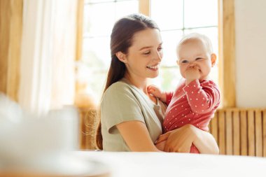 Genç, gülümseyen bir anne kucağında bebeğiyle rahat bir evde oturuyor. Aşk, ilgi, annelik kavramı