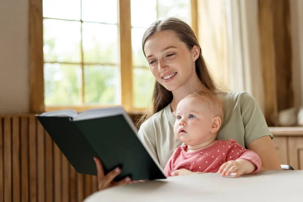 Young beautiful mother reading book for her little cute baby at home. Smiling woman working in kitchen with little child. Concept of remote work and motherhood