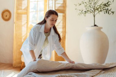 Uyandıktan sonra yatağını toplayan neşeli genç bir kadının portresi. Dinlenmiş, mutlu bir kadın, evinde yastığını tutuyor ve modern yatak odasında duruyor. Ev işleri ve ev işleri.