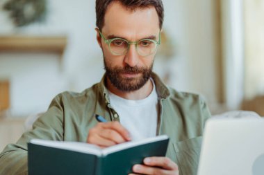Gözlük takan yakışıklı sakallı iş adamı not alıyor, evde ders çalışıyor. Akıllı öğrenci öğrenme dili, online eğitim kavramı 