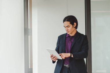 Düşünceli, ciddi Asyalı işadamı dijital tablet kullanıyor, ayakta duruyor, modern ofiste uyukluyor. Siyah takım elbiseli yakışıklı bir Japon, resmi giyinmiş, evden çalışıyor, fotokopi çekiyor. Teknoloji konsepti