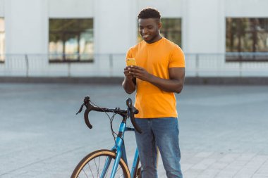 Gülümseyen Afro-Amerikan bir adam cep telefonuyla ekranda bisikletin yanında duruyor. Genç mutlu hippi açık havada mobil uygulama alışverişi yapıyor. Teknoloji konsepti 